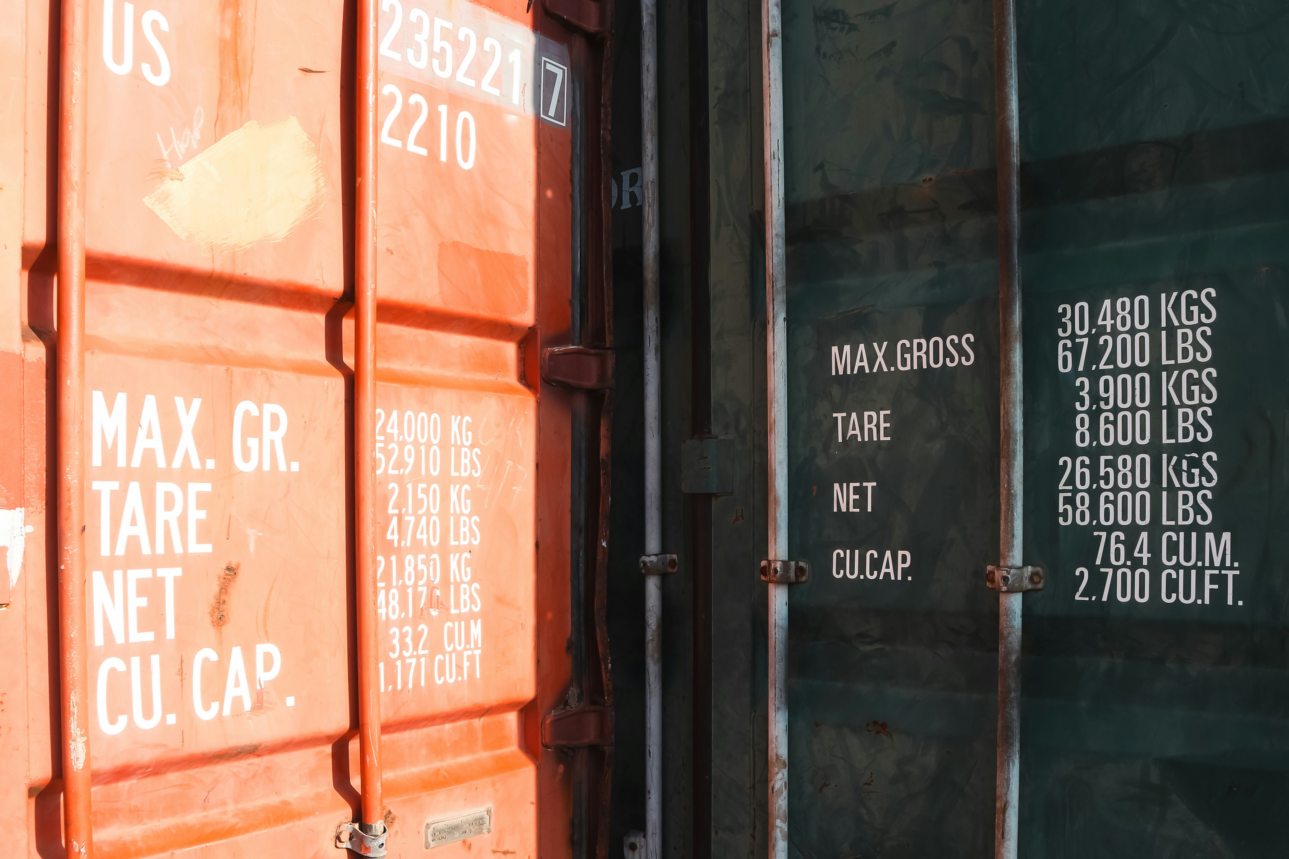 Close-up detail of two shipping containers showing weight markings, max gross, tare, net, and cubic capacity in orange and dark green