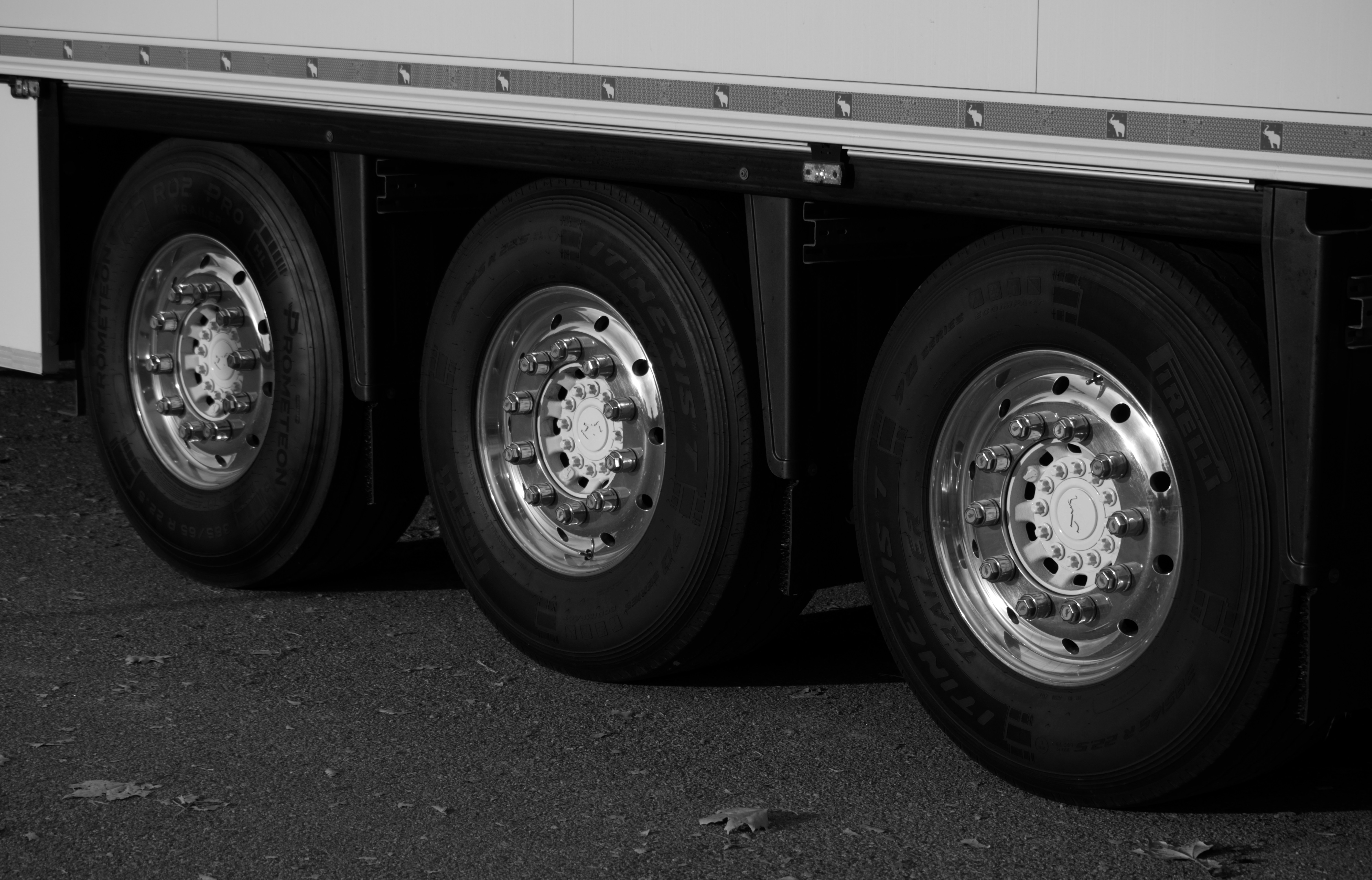 Close-up black and white photograph of three chrome wheels on a heavy truck trailer axle, polished hub caps reflecting light against dark asphalt.