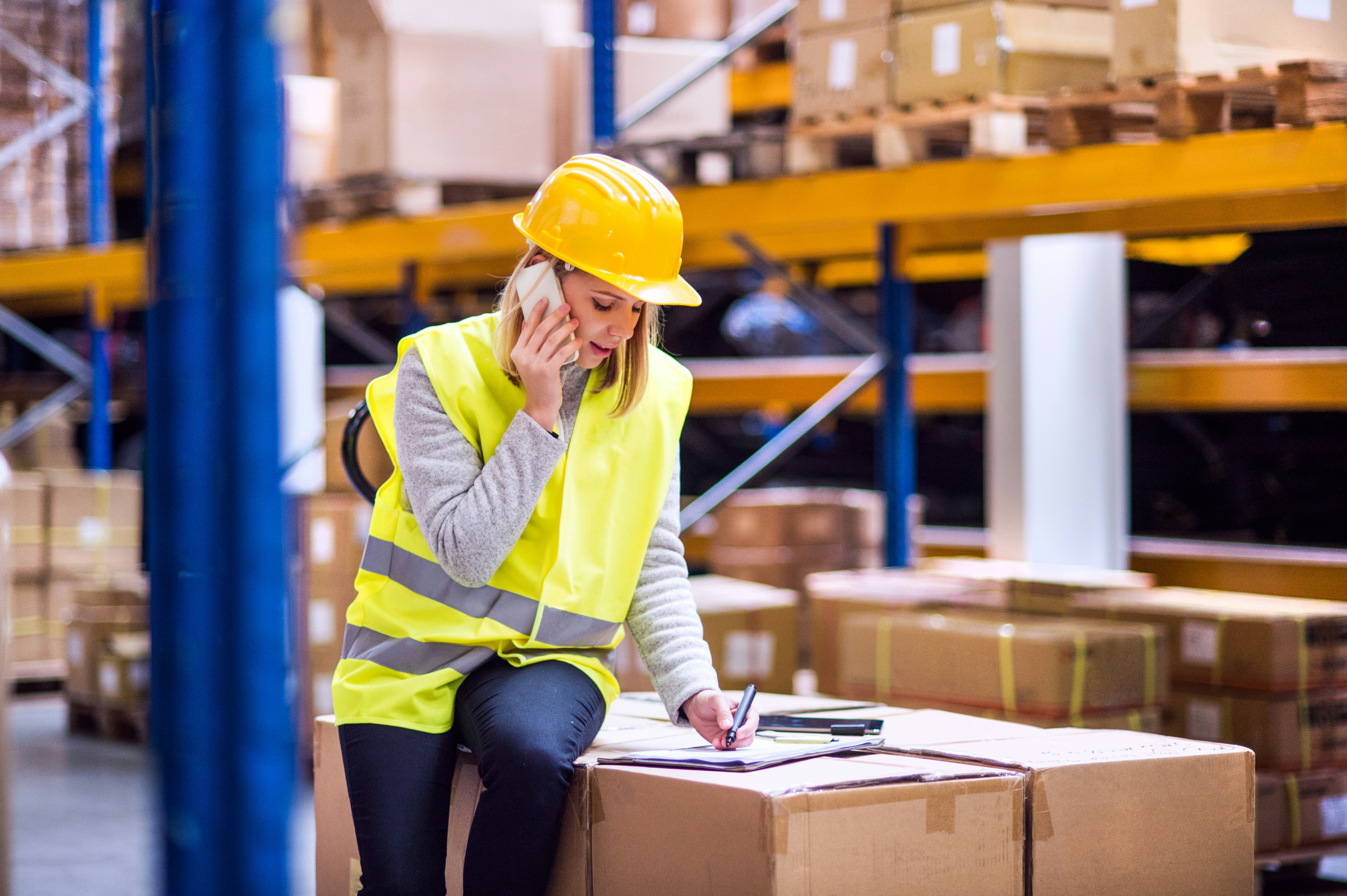 Warehouse worker in a hard hat making a phone call and taking notes