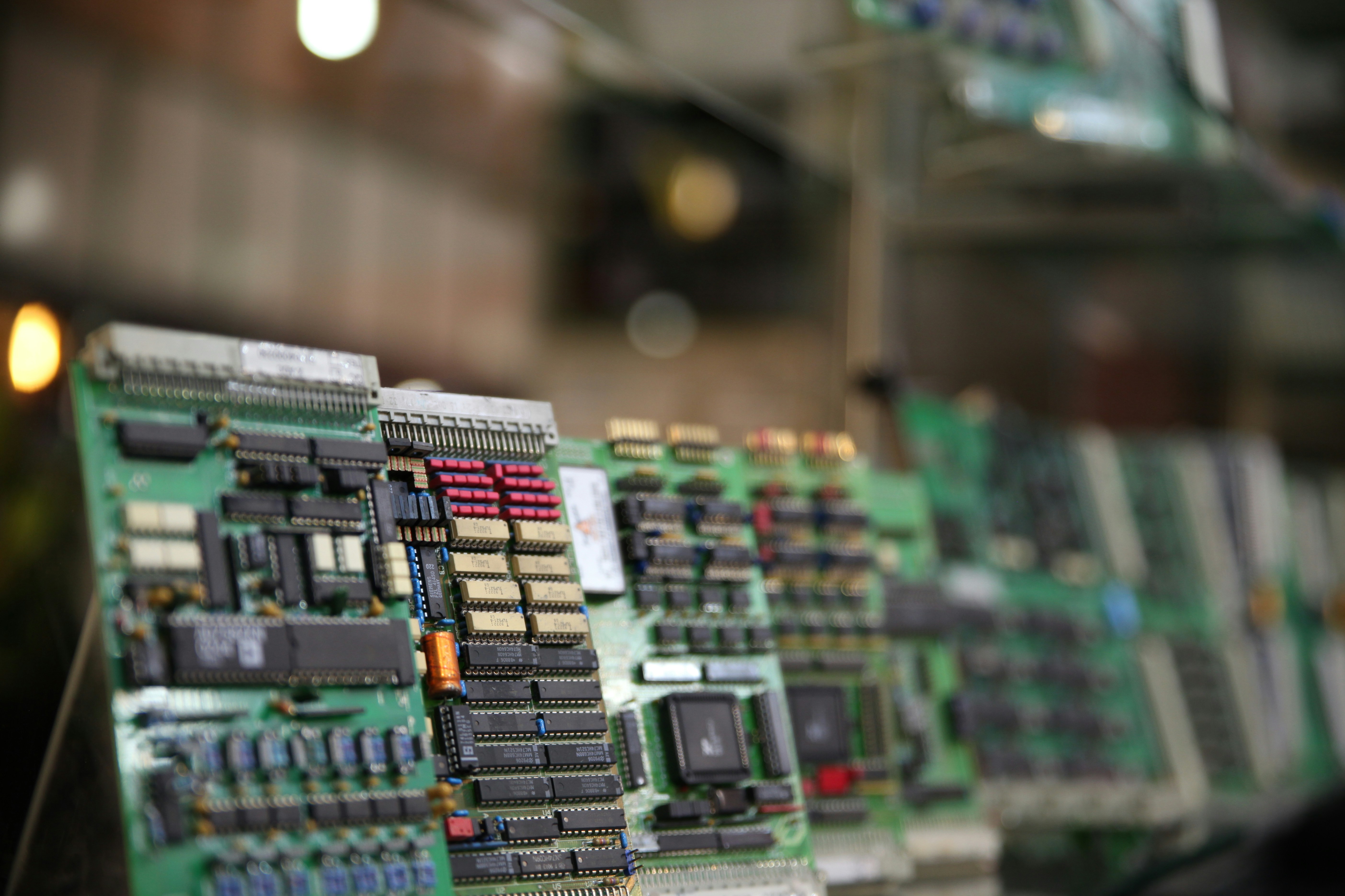 A row of green printed circuit boards stand vertically on a rack in a dimly lit electronics factory, with warm bokeh highlights in the background.