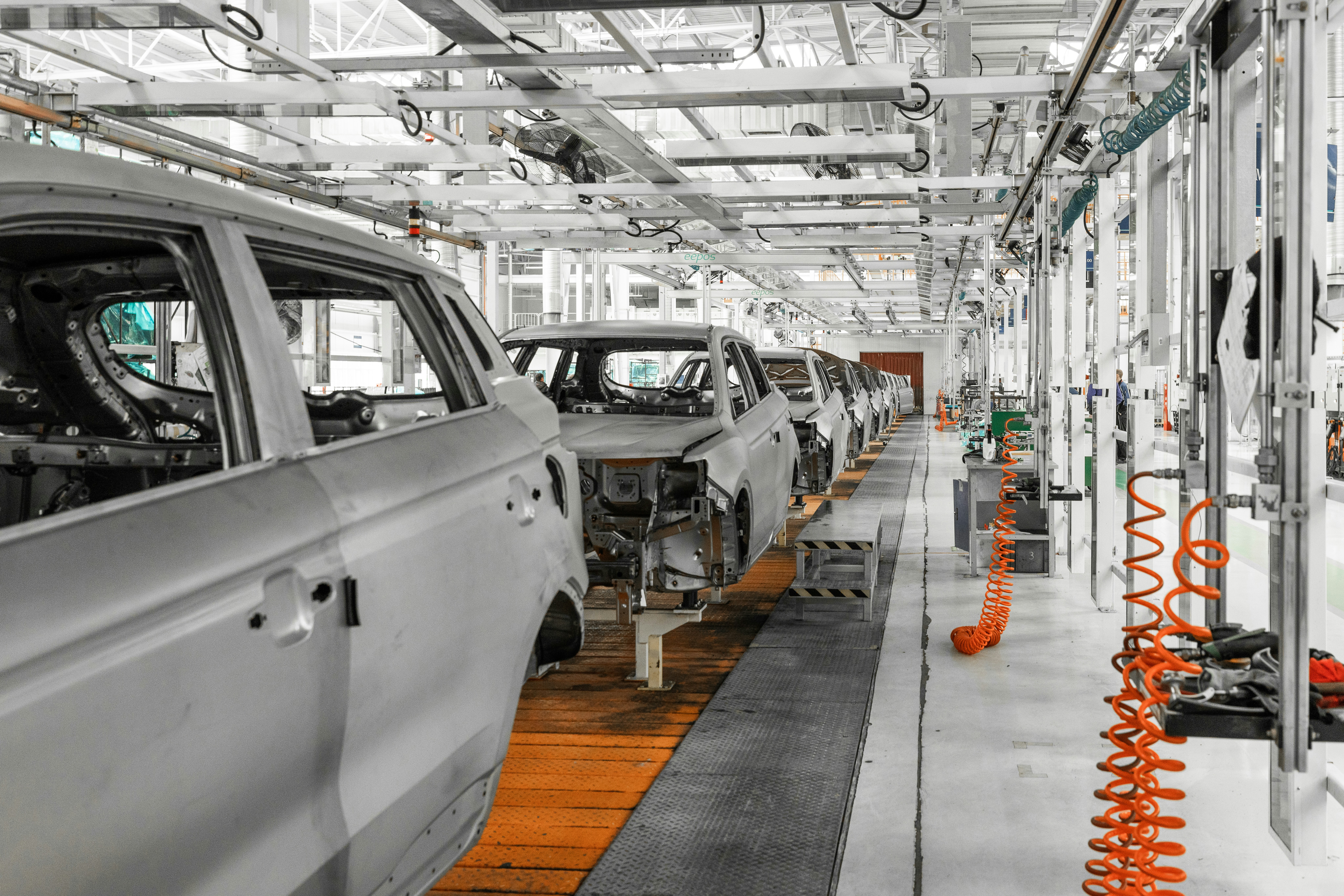Automotive assembly line with silver SUV body shells progressing through production stations, orange coiled air hoses hanging from overhead rail system, clean bright factory floor with tool stations on the right.