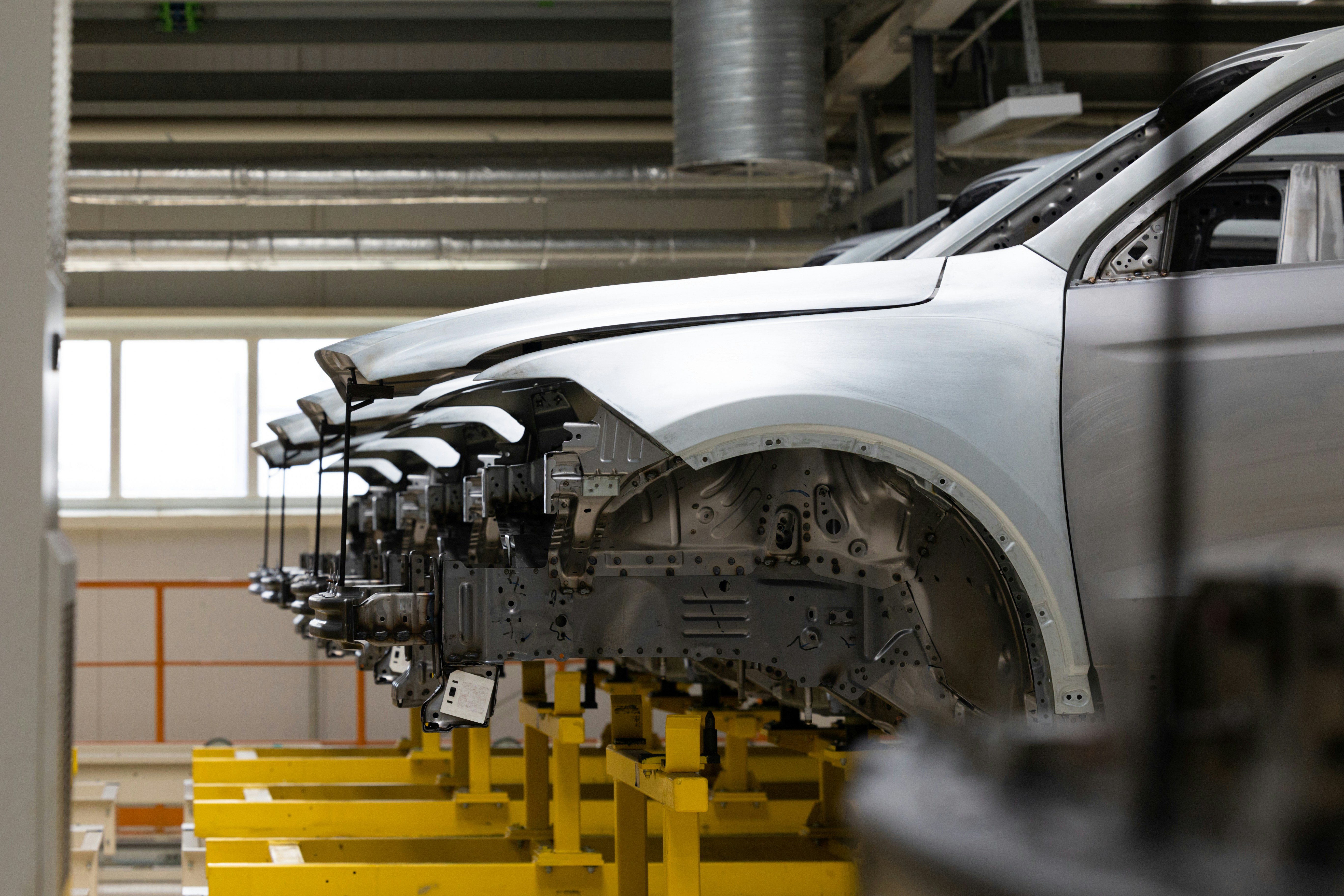 Close-up of silver car body shell on yellow assembly jig in automotive factory, showing front fender and wheel arch area with exposed chassis components and mechanical fixtures beneath.