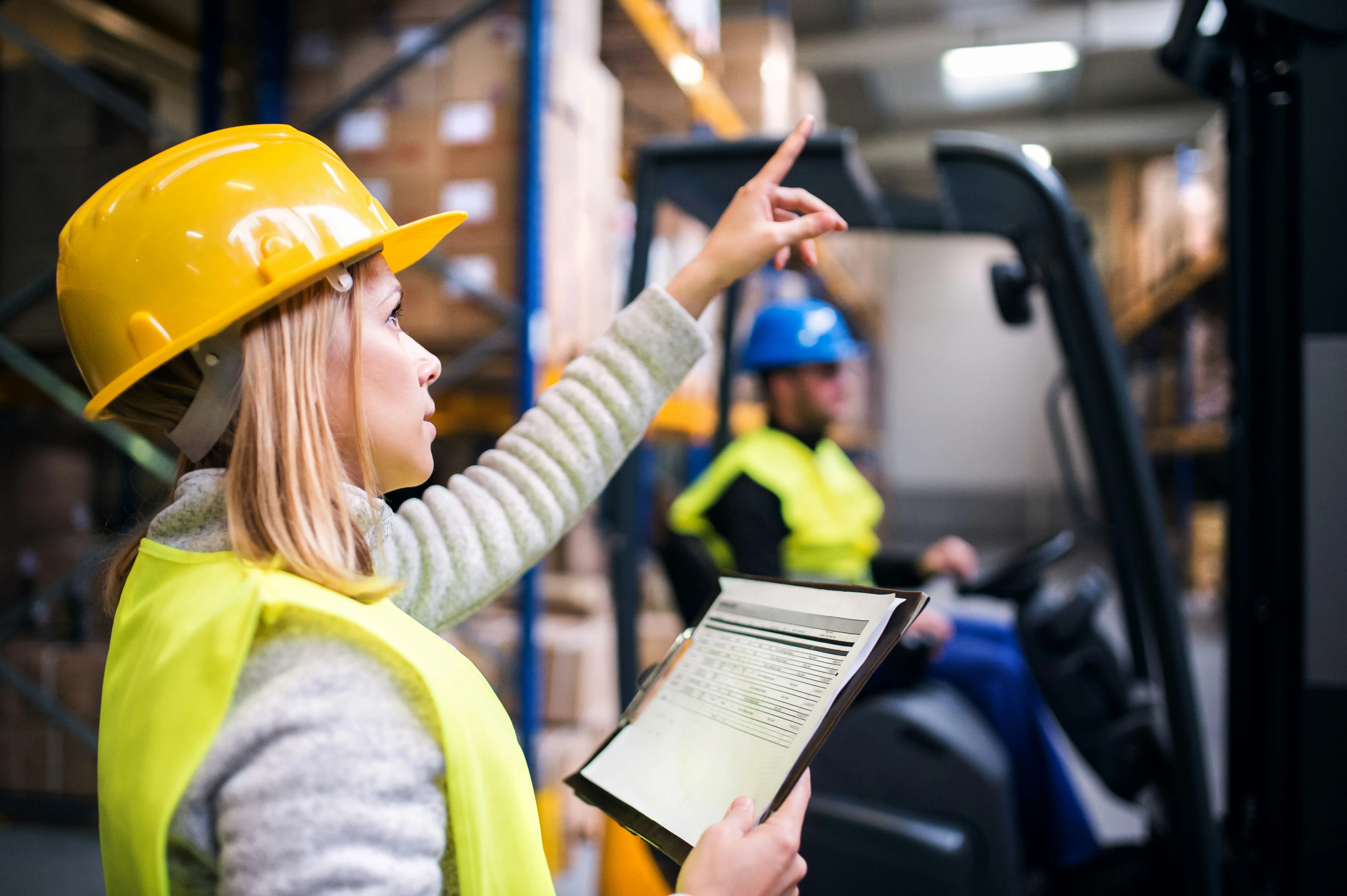 Female warehouse supervisor in hard hat pointing upward while holding clipboard
