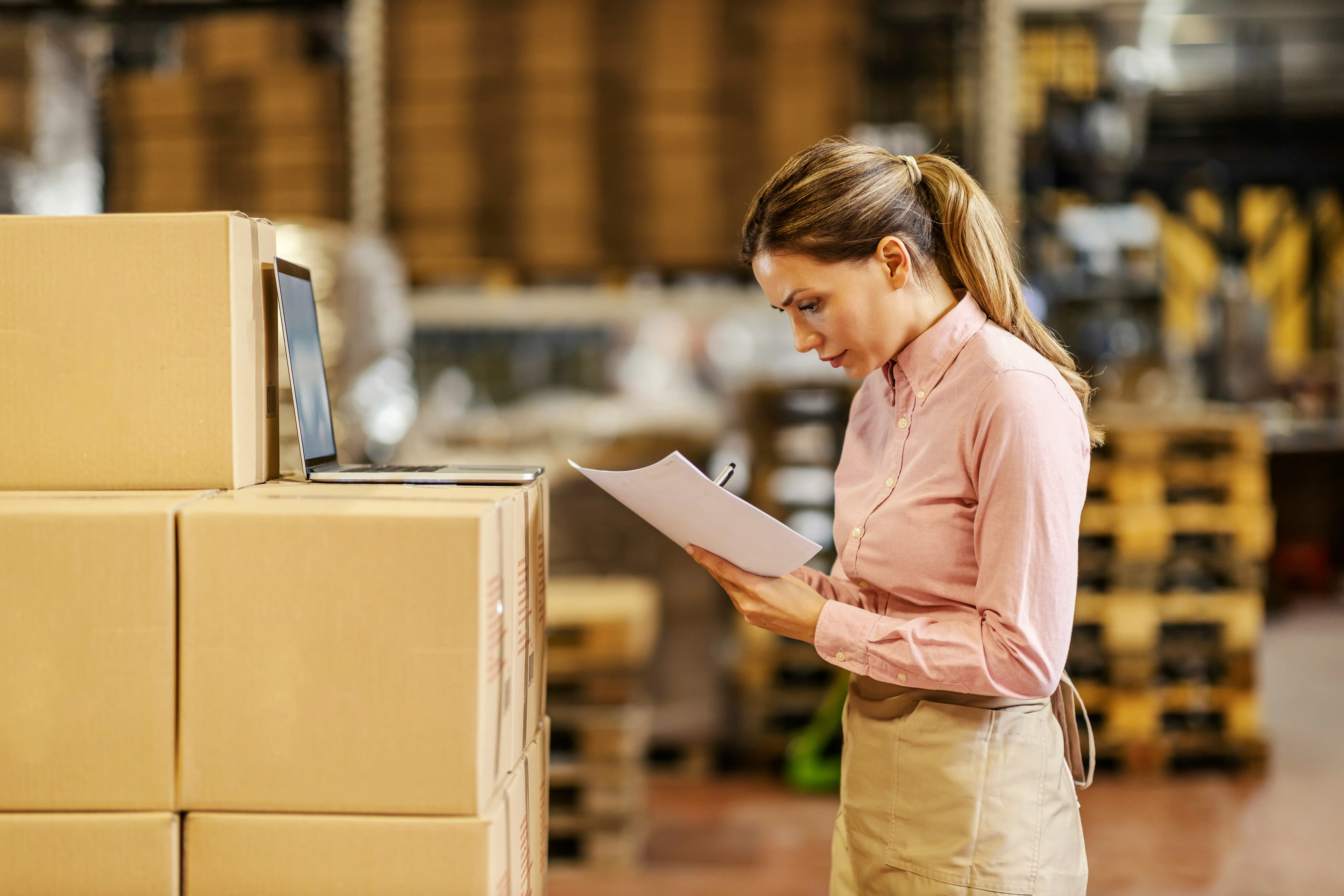 Worker reviewing paperwork beside stacked cardboard boxes with a laptop on top — the manual reality of spreadsheets, exports, and partner templates