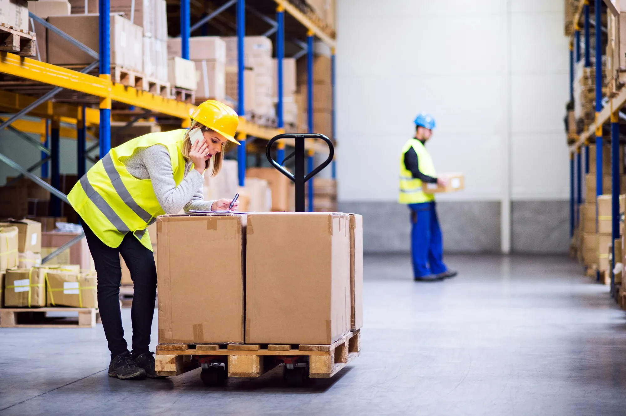 Warehouse worker making a phone call and taking notes beside a pallet jack.